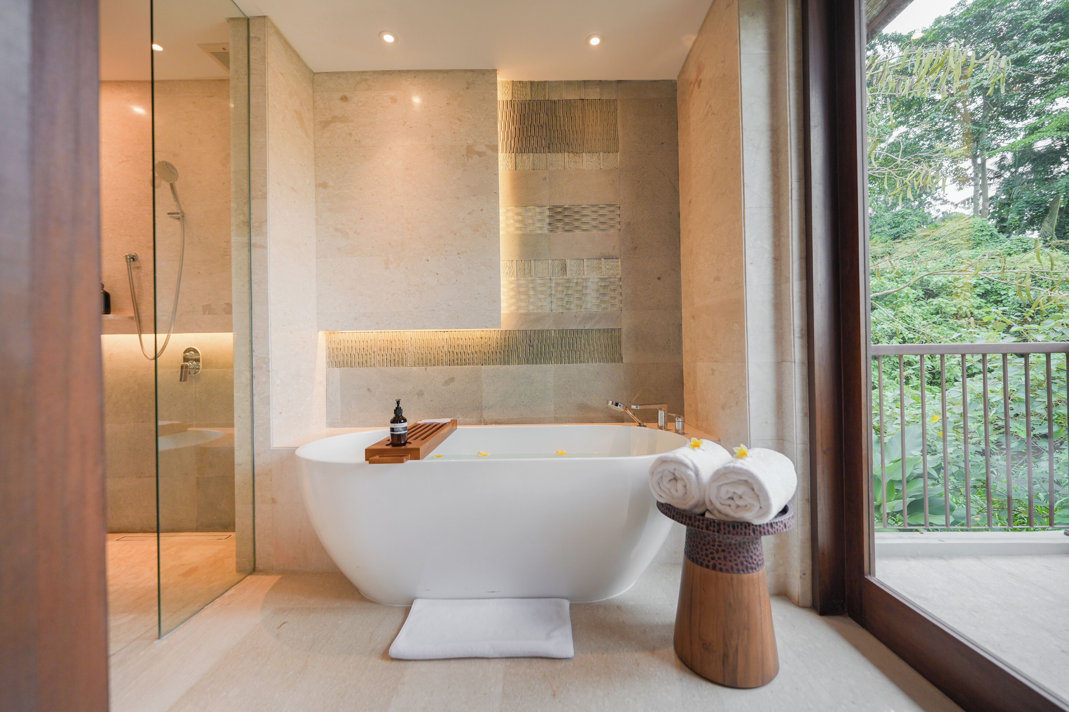 A modern bathroom with a freestanding bathtub, a glass shower enclosure, and a view of greenery through a large window.