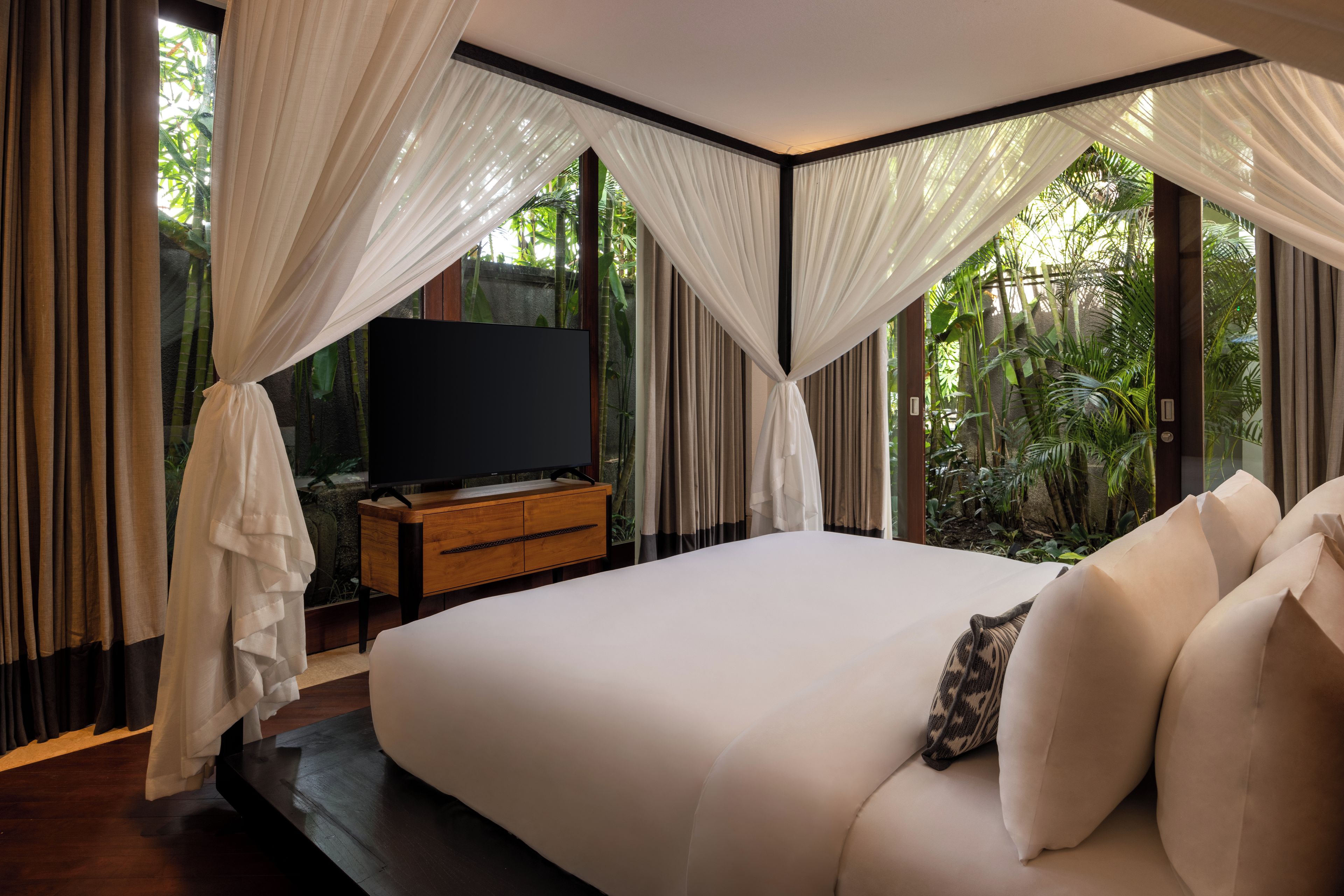A bedroom with a large bed, a TV on a wooden cabinet, and a view of greenery through the windows.