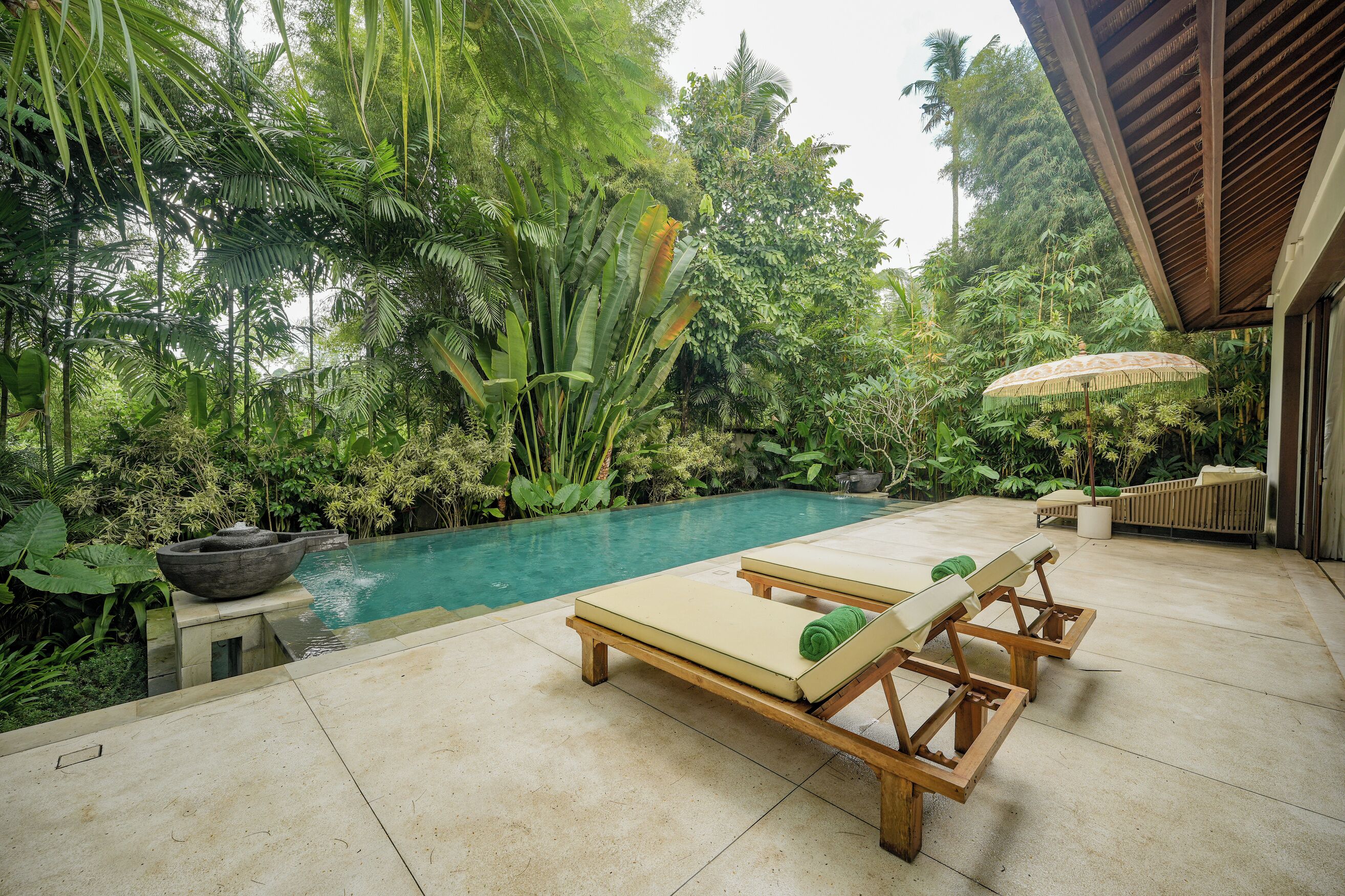 A poolside area with lounge chairs, a stone basin, and lush tropical vegetation.
