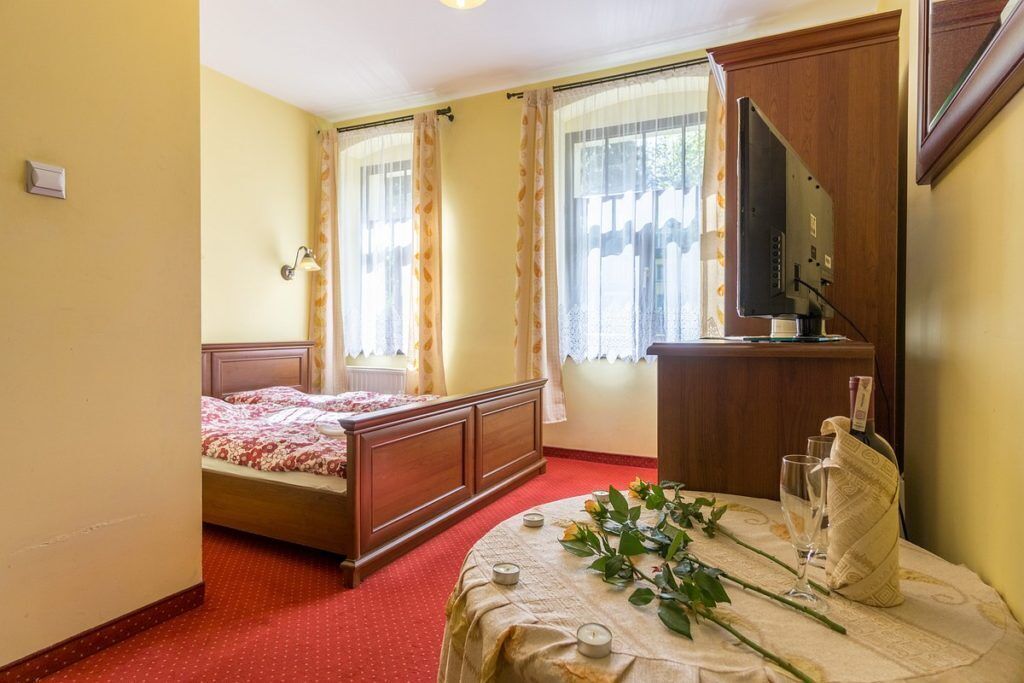 Un dormitorio con cama, una mesita de noche con una botella y vasos, un televisor y una ventana con cortinas.