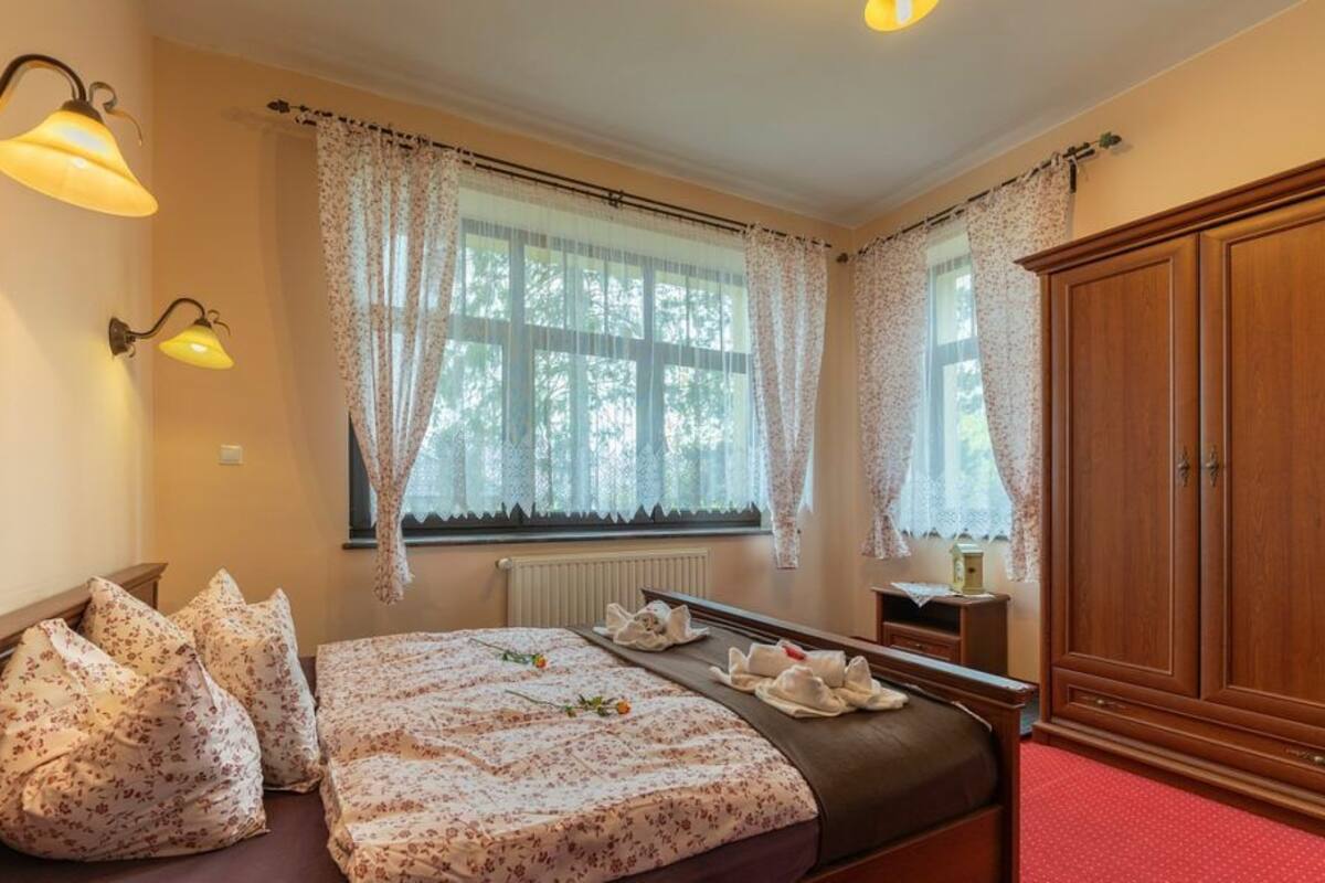 Un dormitorio con cama, mesitas de noche, armario y dos ventanas con cortinas.