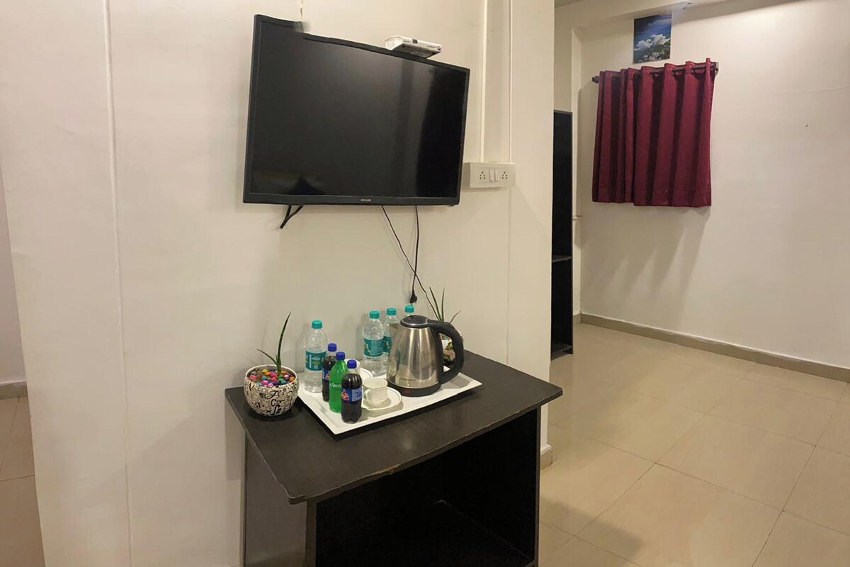 A room with a flat-screen TV mounted on the wall, a small table with a kettle, bottles, and a decorative bowl.