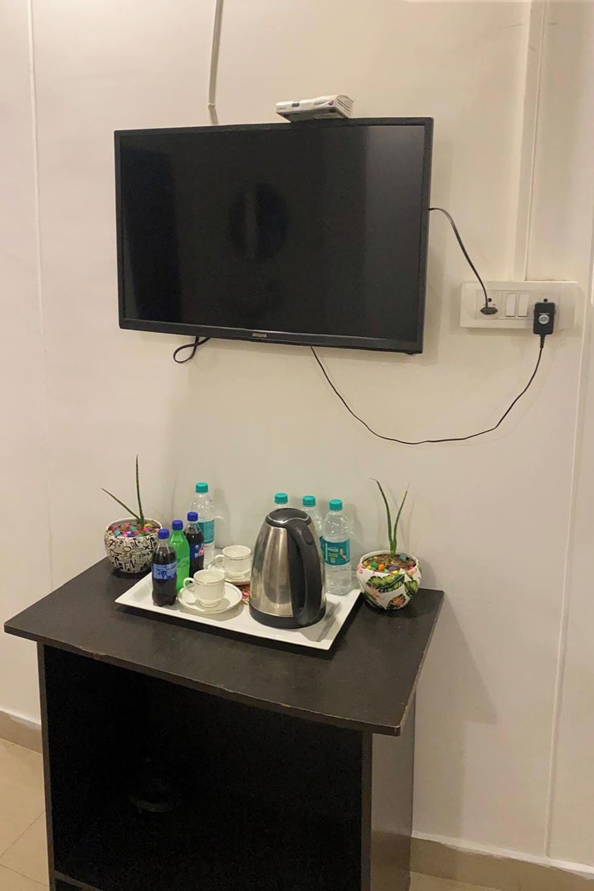 A wall-mounted TV above a small table with a tray containing a water bottle, a cup, and a thermos.