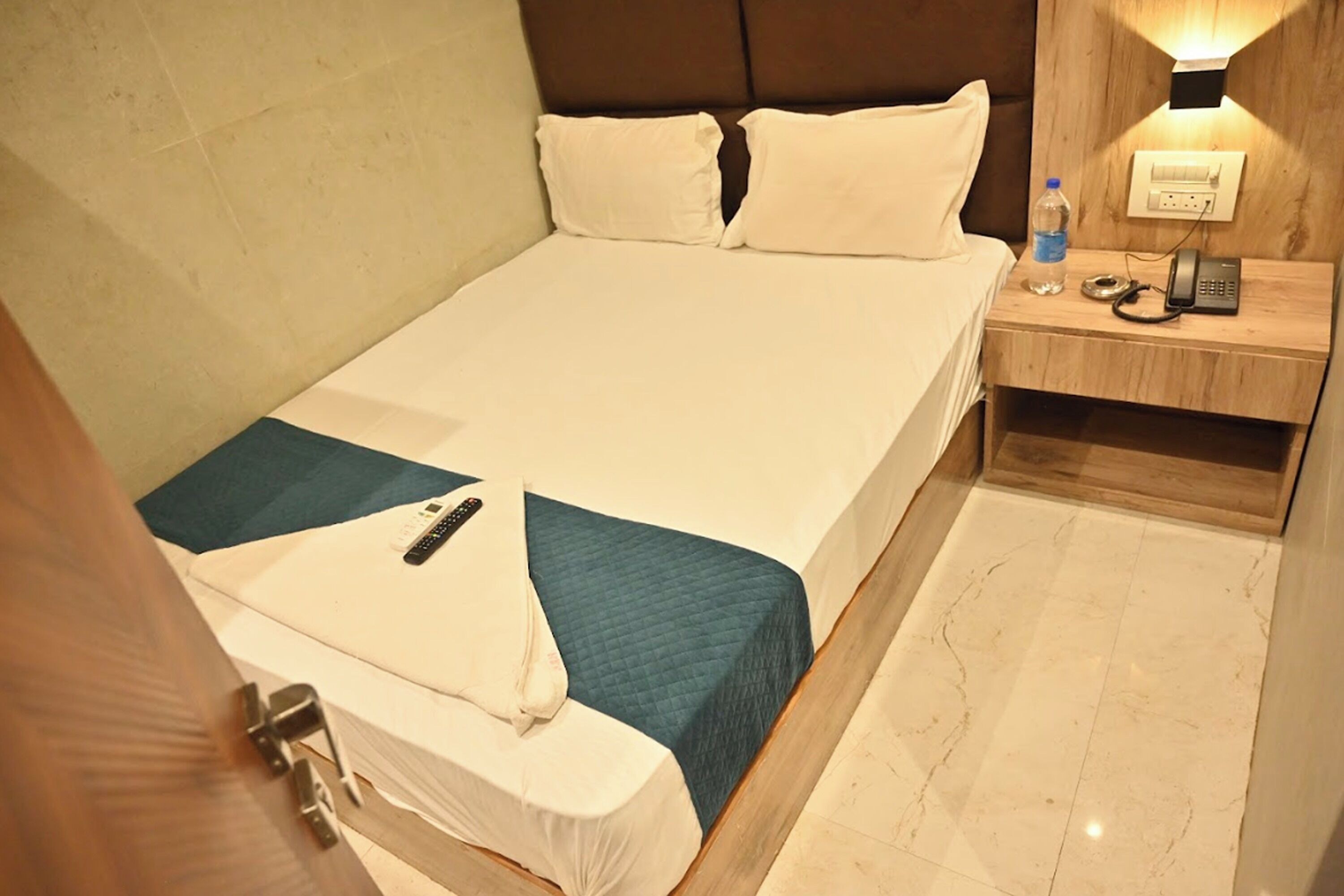A hotel room with a large bed, a bedside table with a phone and a bottle, and a wall-mounted light.