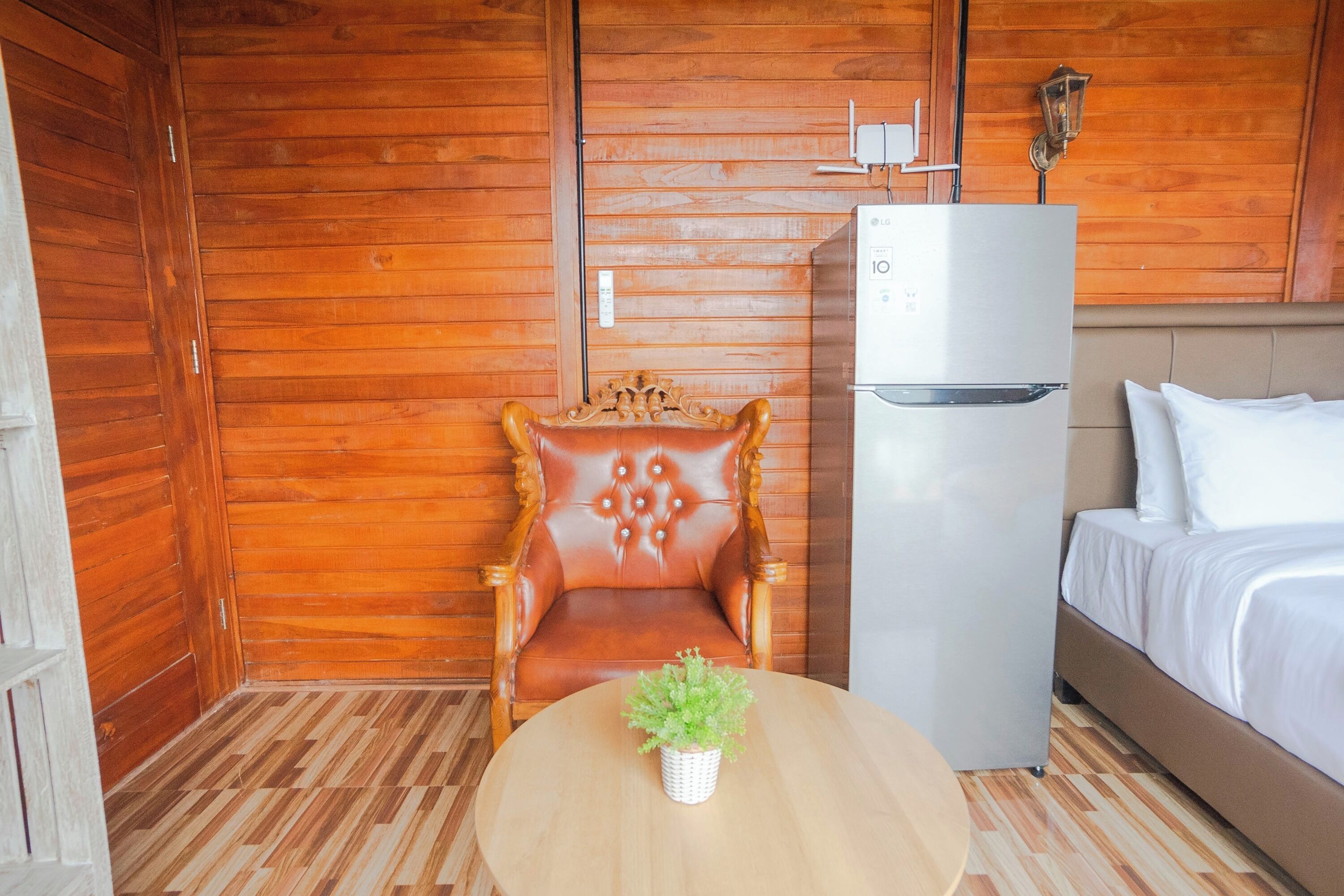 A room with wooden paneling, a bed, a white refrigerator, a wooden chair, and a small table with a plant.
