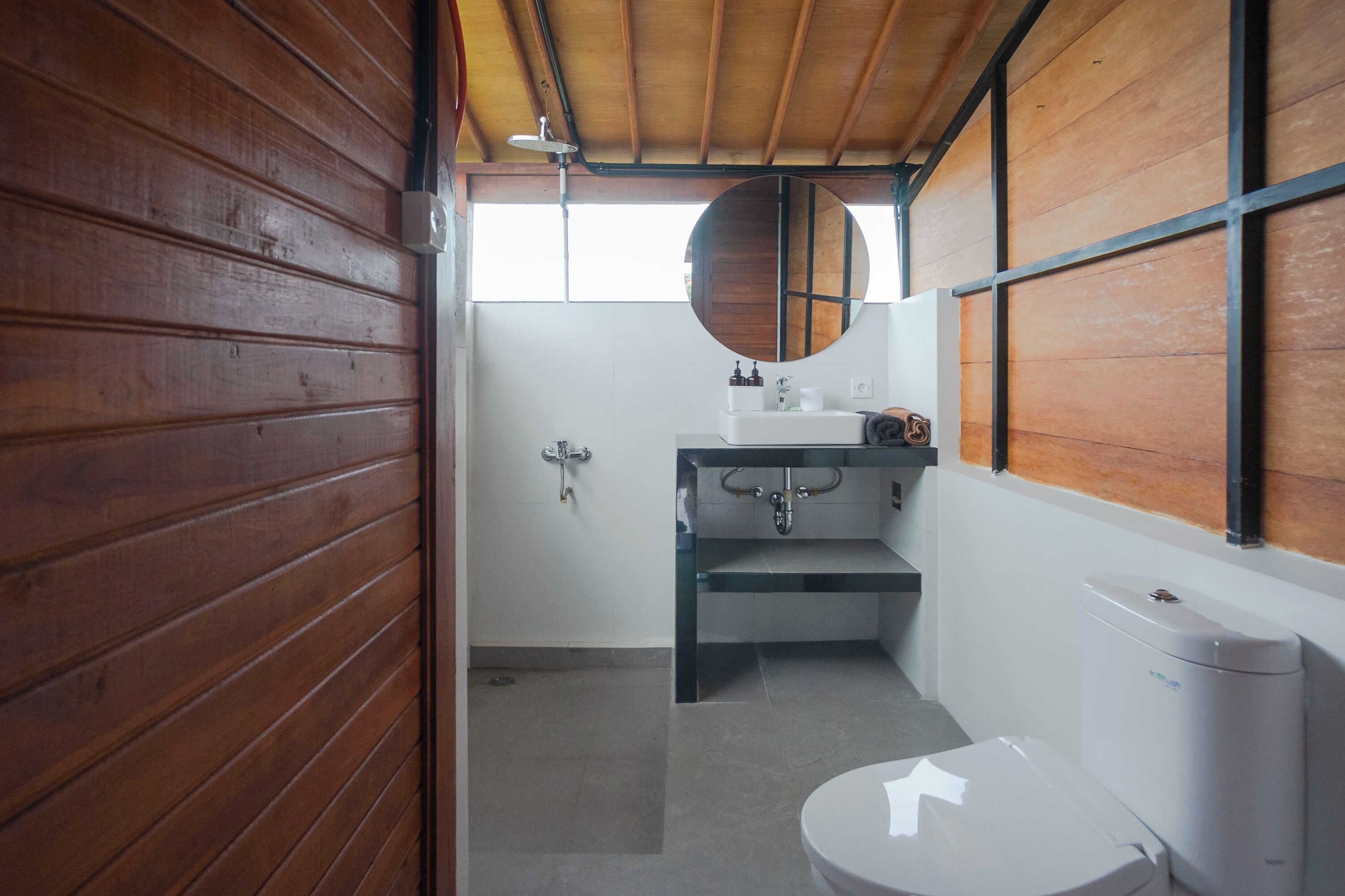 Une petite salle de bain avec un mur en bois, des toilettes blanches, un lavabo avec un miroir au-dessus et un espace douche.