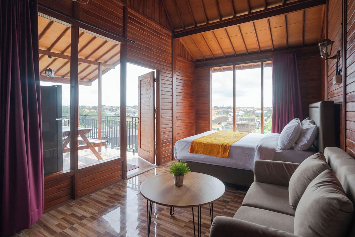 Ein Zimmer mit Holzboden, einem Bett, einer Couch, einem kleinen Tisch mit Pflanze und großen Fenstern mit Blick auf die Stadt.