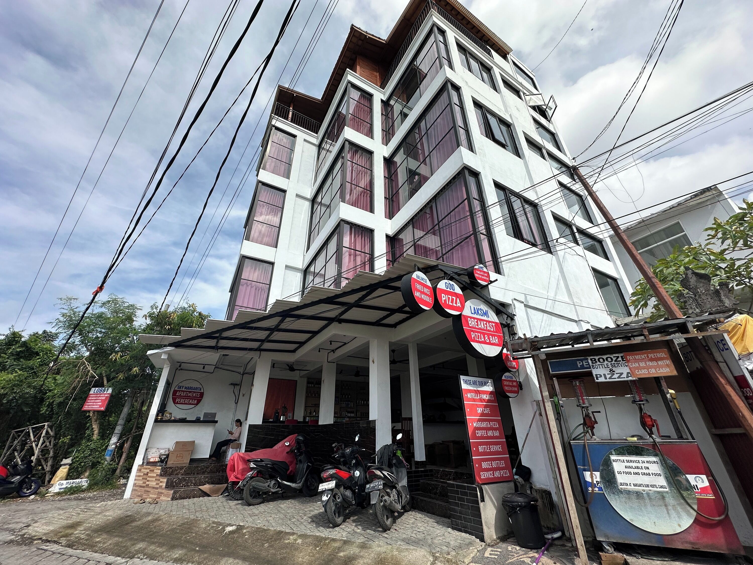 Un edificio de varios pisos con un acceso cubierto y varios letreros que anuncian servicios como pizza y desayuno.