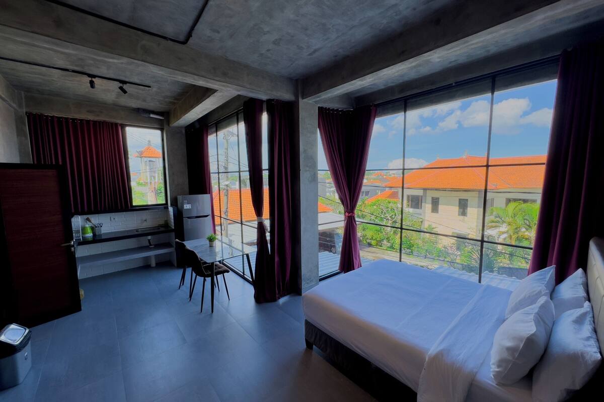 Ein modernes Hotelzimmer mit einem großen Bett, einem Schreibtisch und Blick auf Gebäude durch große Fenster.