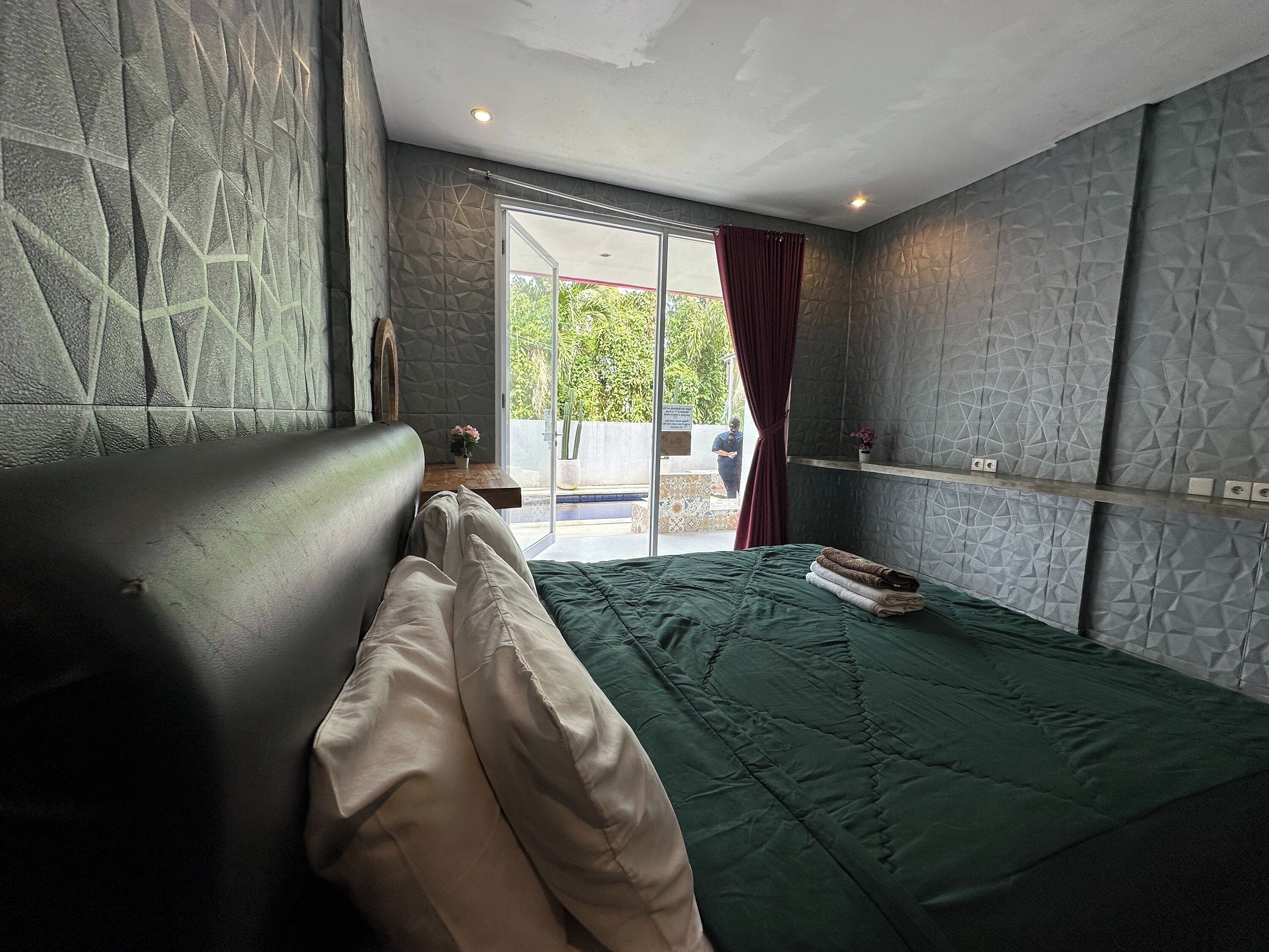 A modern bedroom with a large bed, a small table with a vase, and a view of greenery outside.