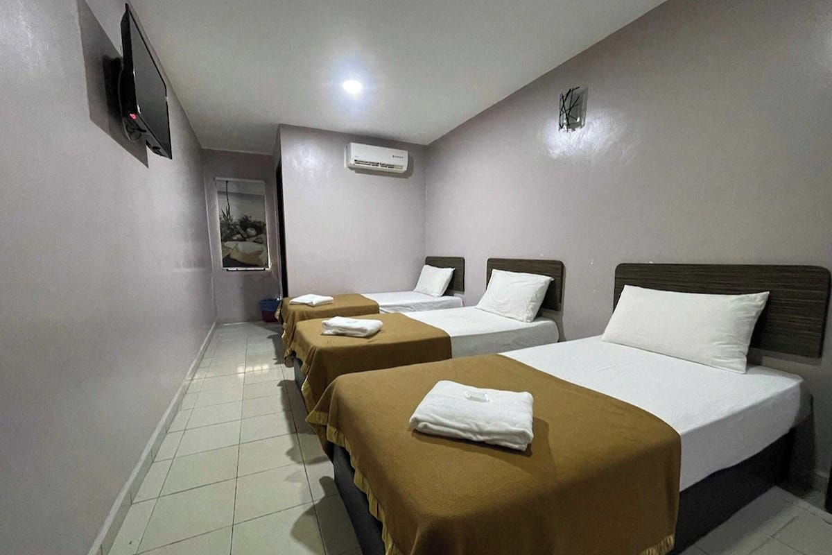 A hotel room with three beds, each with a brown cover and white pillows. There is a flat-screen TV mounted on the wall, an air conditioning unit, and a small window with a view of the outside.