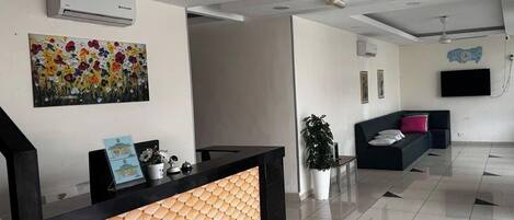 Un área de recepción moderna con un mostrador negro, un cuadro floral de colores, una planta en maceta, un sofá negro con un cojín rosa y un televisor de pantalla plana montado en la pared.