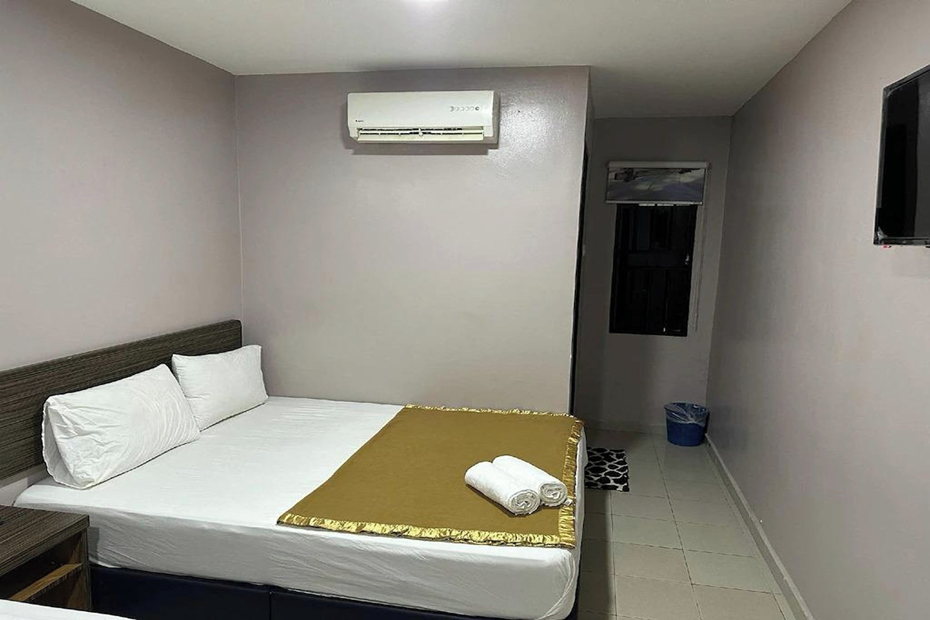 A hotel room with a bed, a wall-mounted air conditioner, a window, and a trash bin.