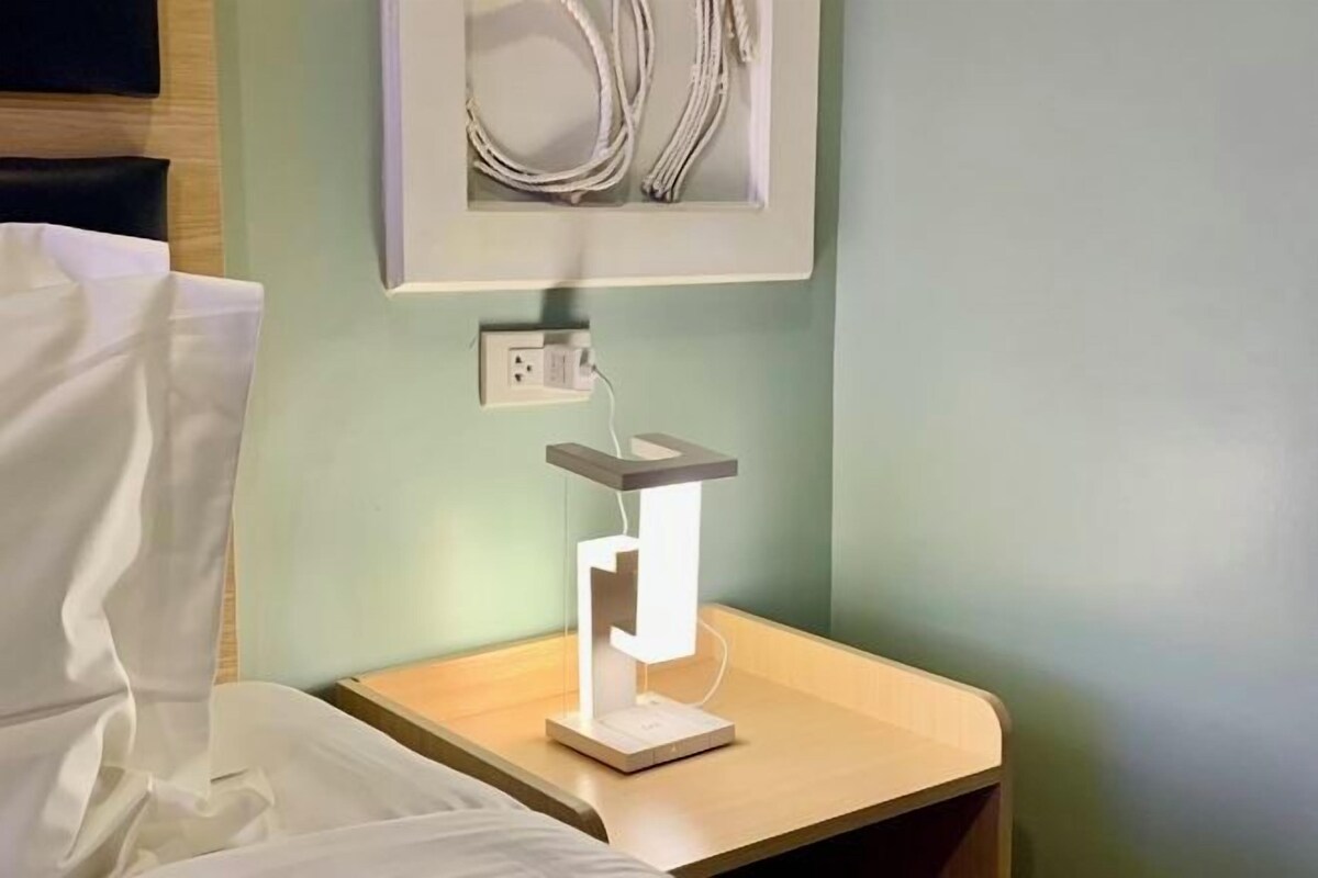 A bedside table with a modern lamp, a power outlet, and a framed picture on the wall.