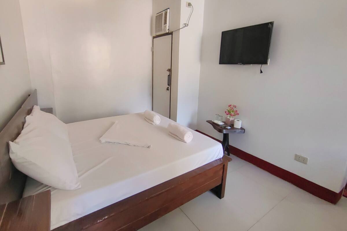 A neatly made bed with white linens and a rolled towel, a small bedside table with a vase of flowers, a wall-mounted TV, and a door with a mirror.
