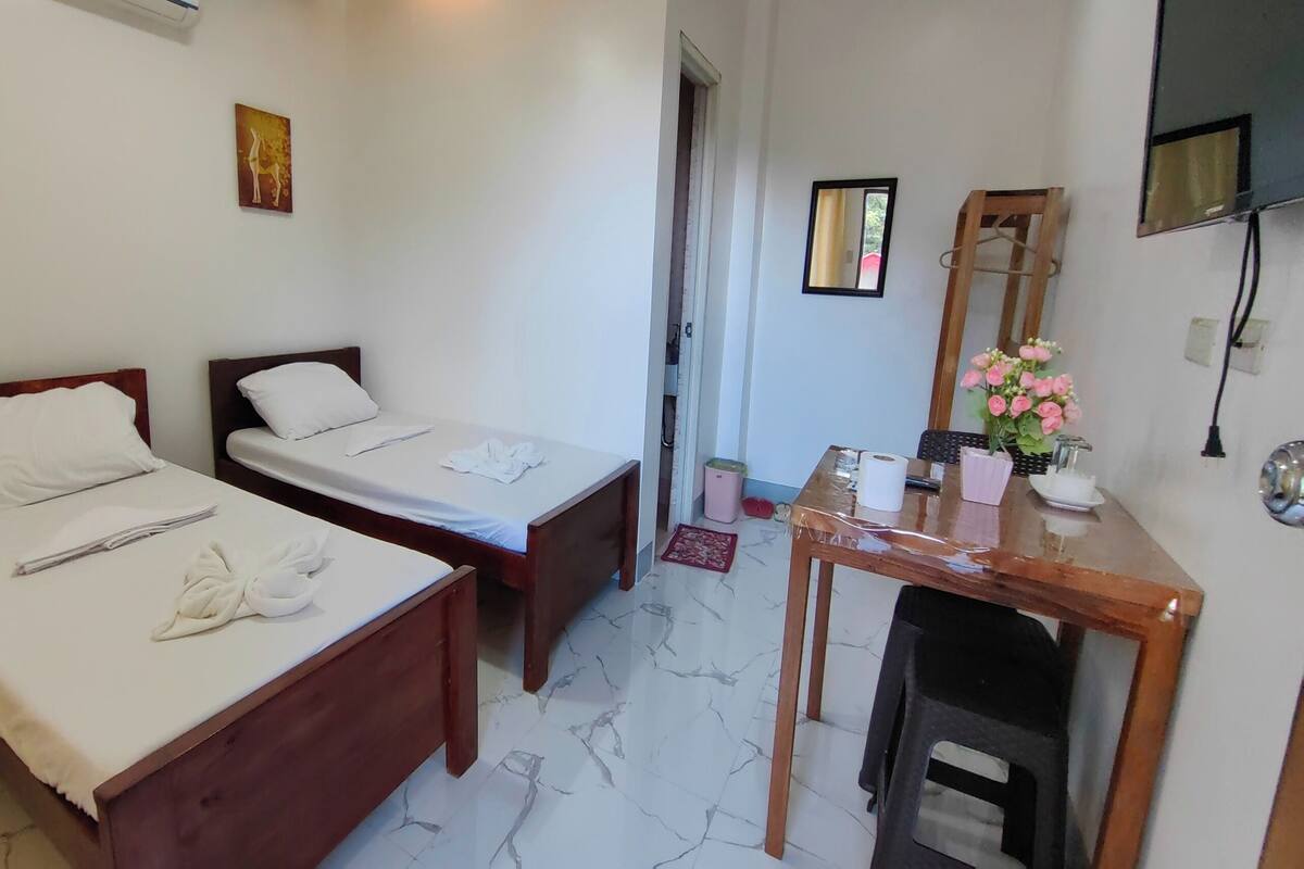 A hotel room with two single beds, a wooden desk with a mirror, a small table with a vase of flowers, and a TV mounted on the wall.