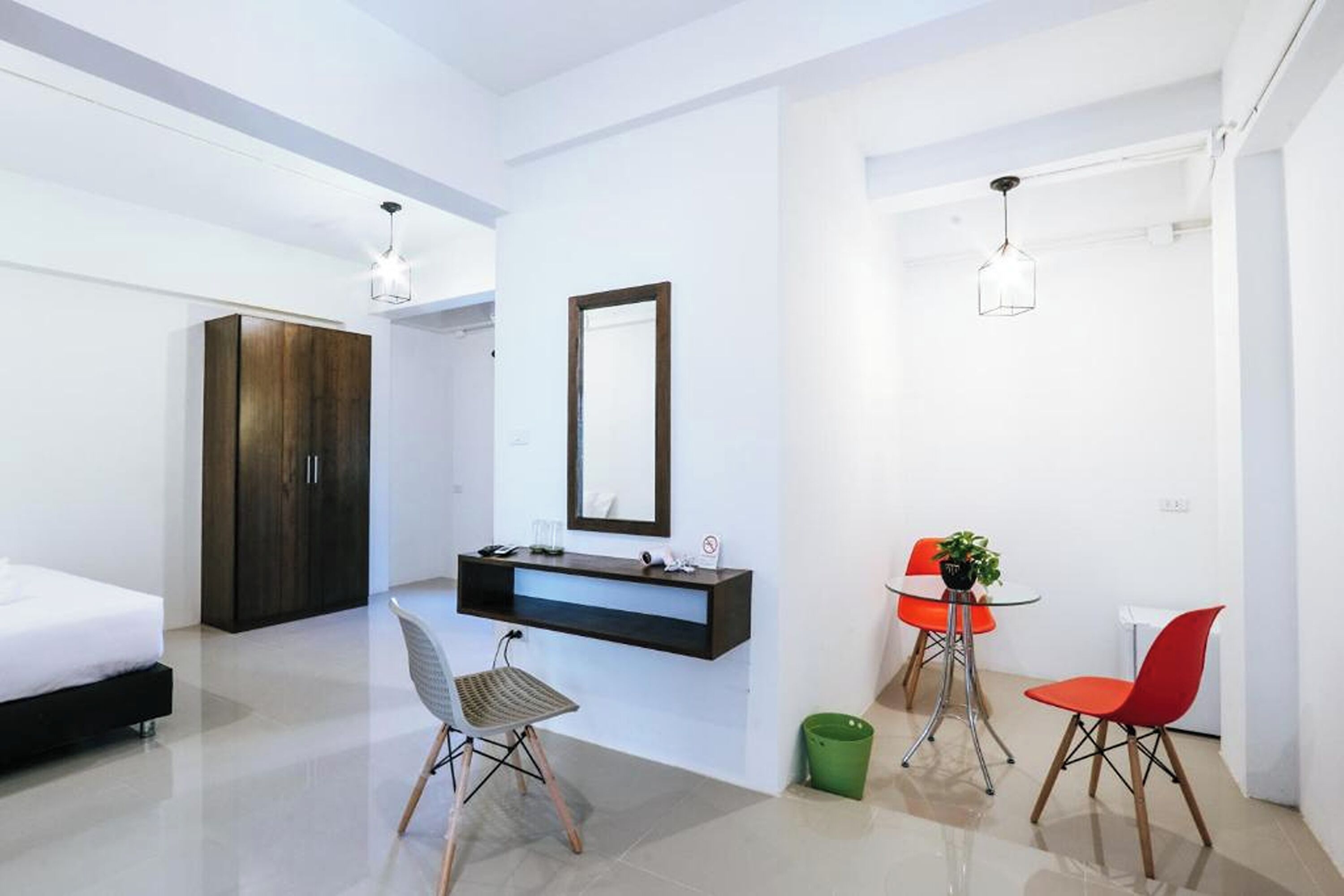 A modern bedroom with a bed, a desk, and a chair. There is a mirror on the wall and a green trash bin on the floor.