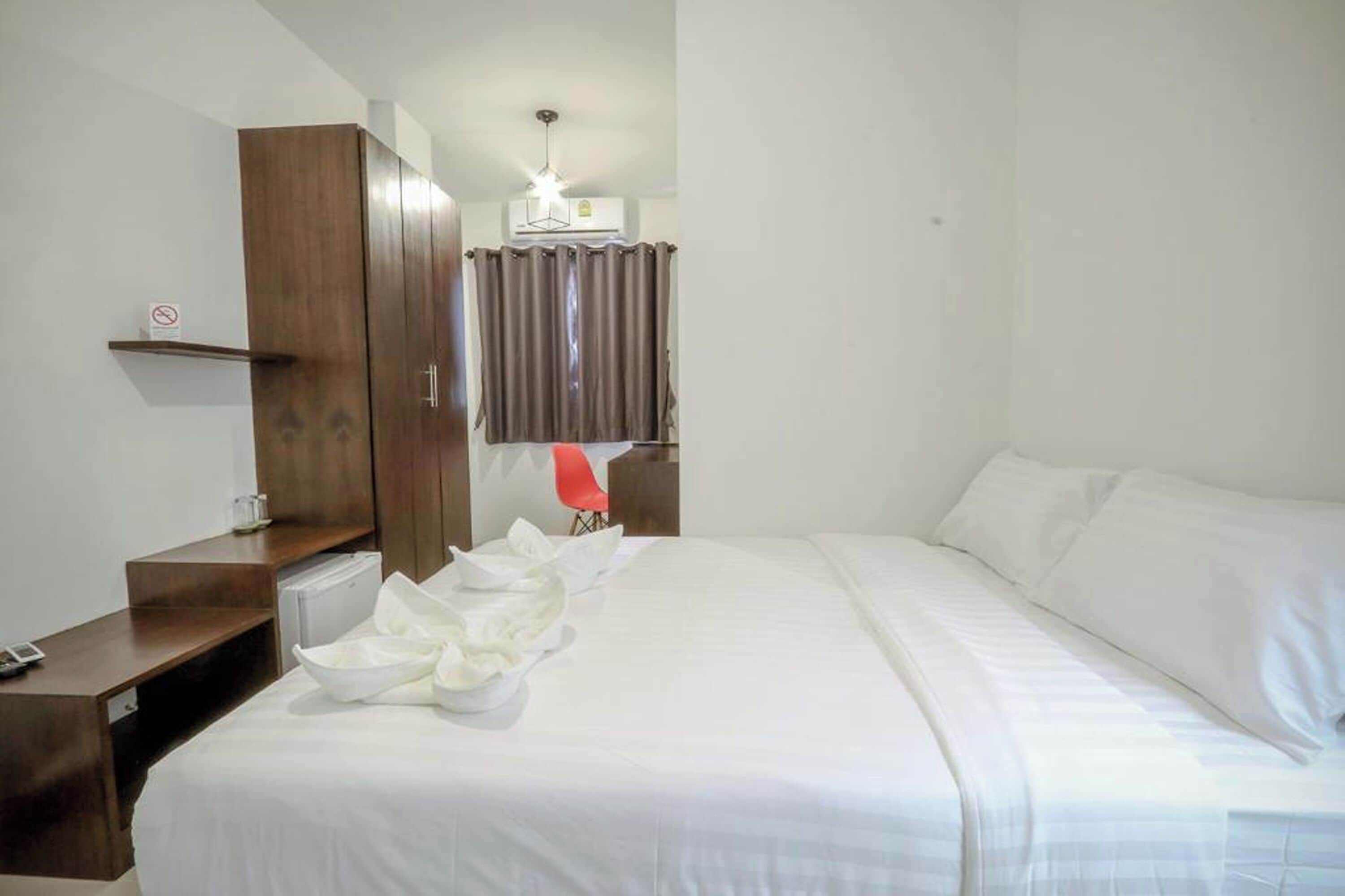 A hotel room with a bed, a wooden wardrobe, a desk with a chair, a small table, and a window with curtains.