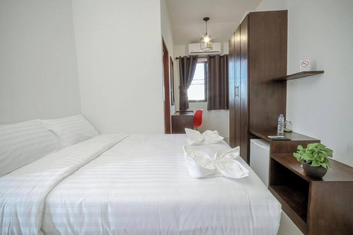 Una cama bien hecha con ropa blanca, una silla roja, un escritorio pequeño con una planta y un aire acondicionado instalado en la pared.