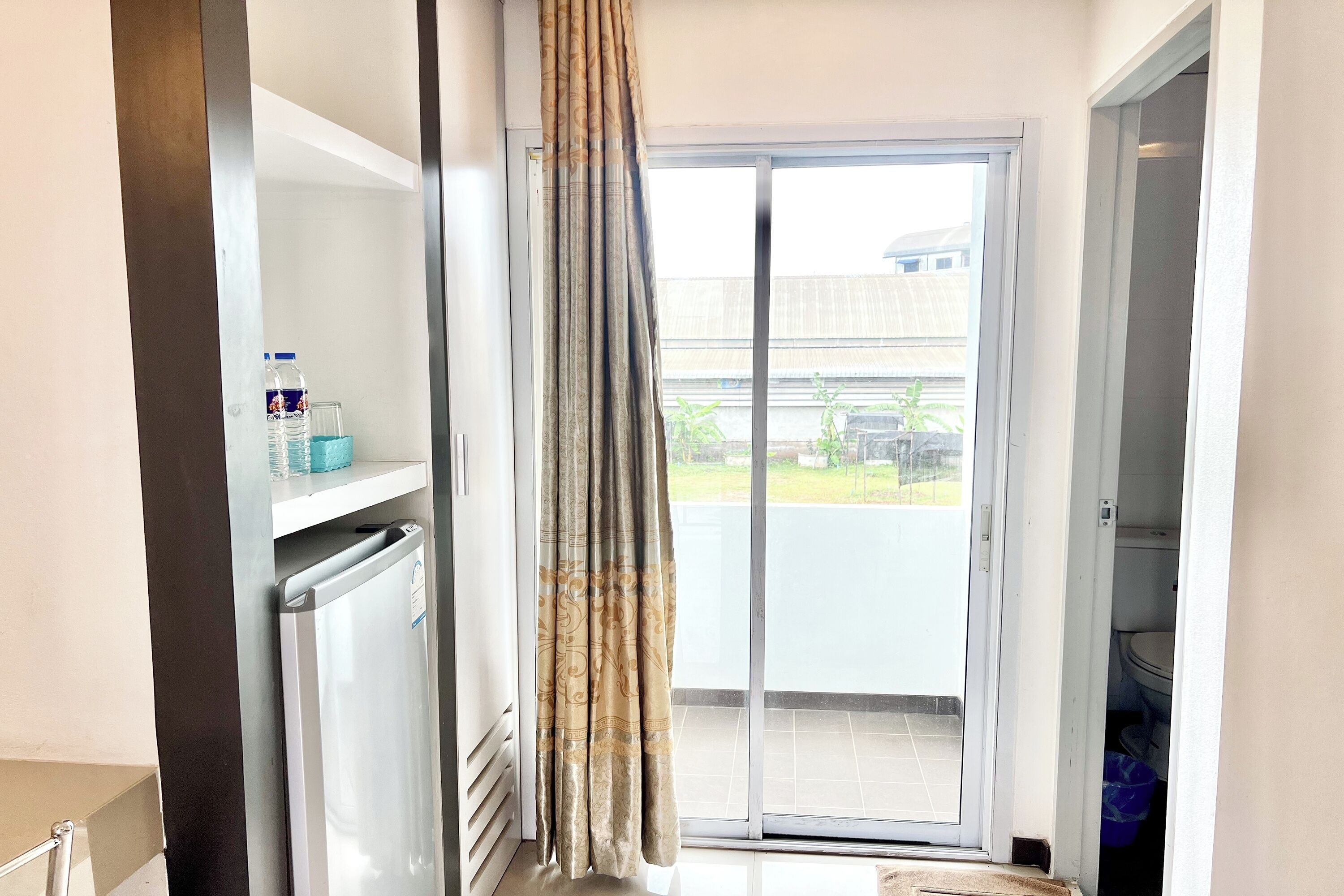 A room with a sliding glass door leading to a balcony, a refrigerator, and a shelf with a water bottle and glass.