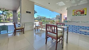 A hotel lobby with a tiled floor, wooden chairs, a reception desk, and a large window offering a view of the outside. - Grand Place Resort (Udon Thani)