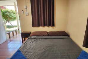 Un dormitorio con una cama, dos almohadas marrones, una colcha gris, una cortina marrón, una mesita de noche y un enchufe de pared.