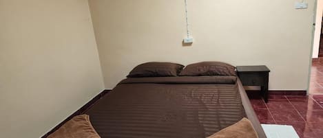 Una cama individual con un colchón oscuro y dos almohadas, una mesita de noche pequeña y un enchufe en la pared.