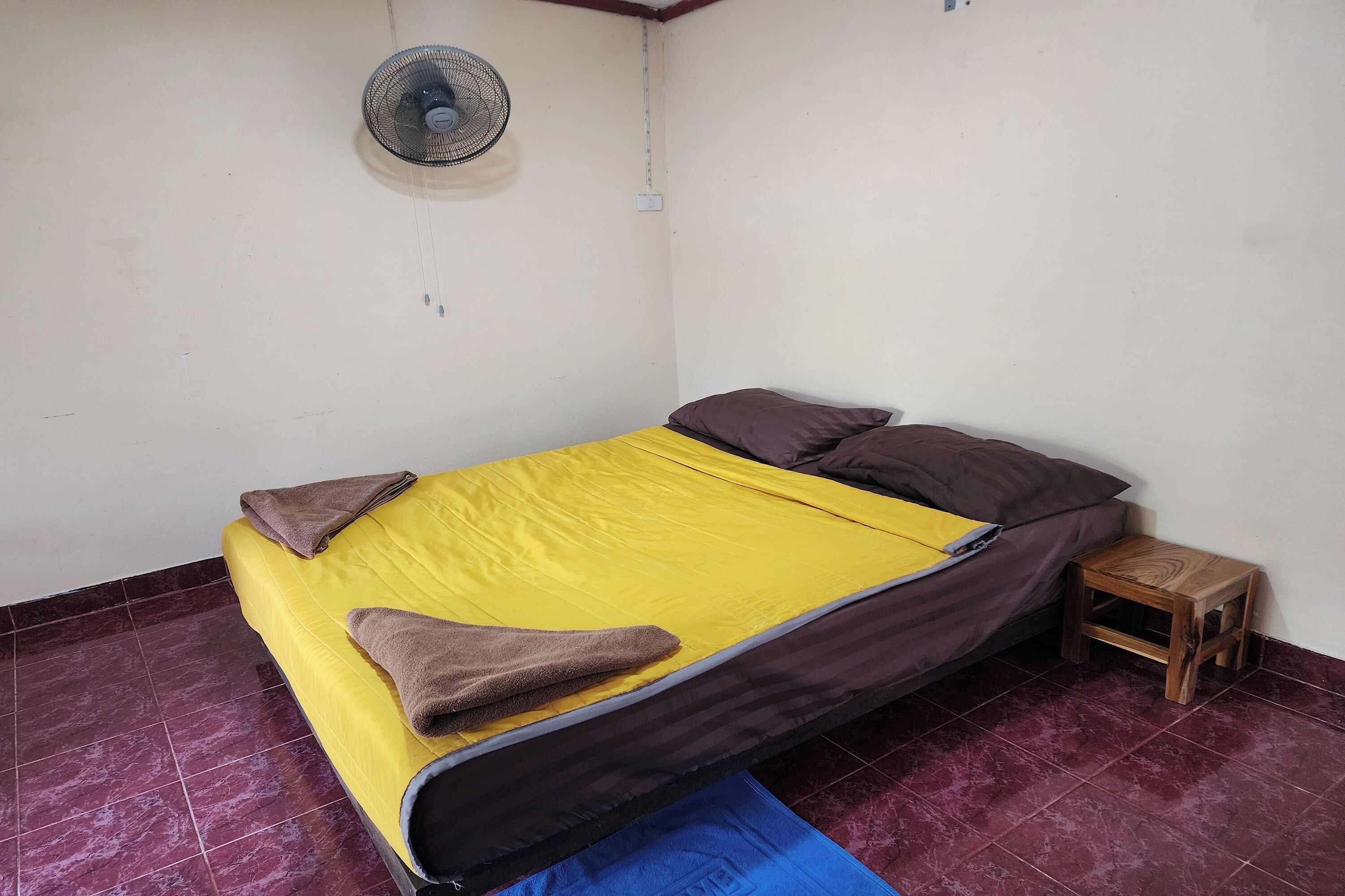 A single bed with a yellow and brown bedspread, two pillows, a wooden nightstand, and a wall-mounted fan.
