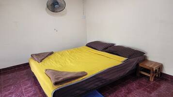A single bed with a yellow and brown bedspread, two pillows, a wooden nightstand, and a wall-mounted fan.