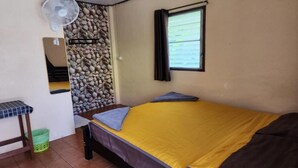 Une petite pièce comprenant un lit, un ventilateur, une fenêtre et un mur décoré de pierres.