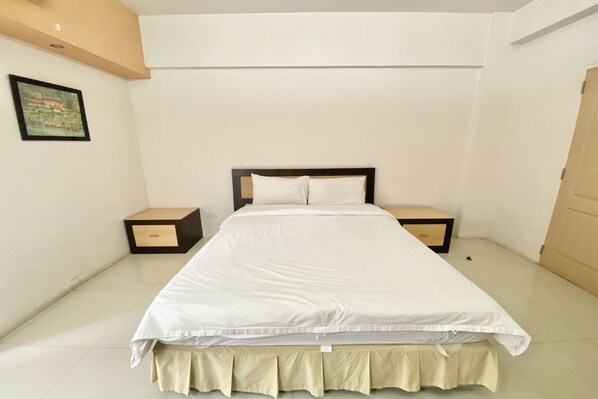 Una cama bien hecha con una colcha blanca y almohadones a juego, flanqueada por dos mesitas de noche con cajones.
