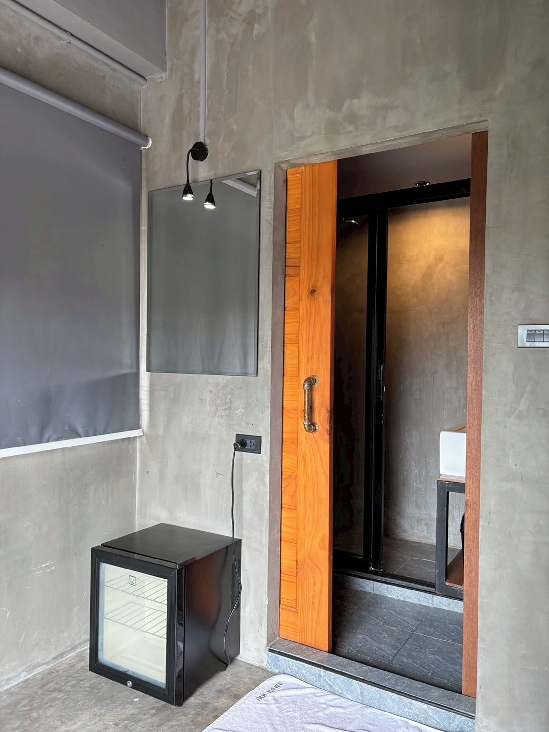 A modern room with a concrete wall, a wooden door, a small black refrigerator, and a ceiling light fixture.