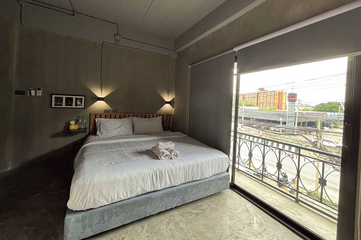 A bedroom with a bed, bedside lamps, a window with a view of a train and buildings, and a small shelf with items.