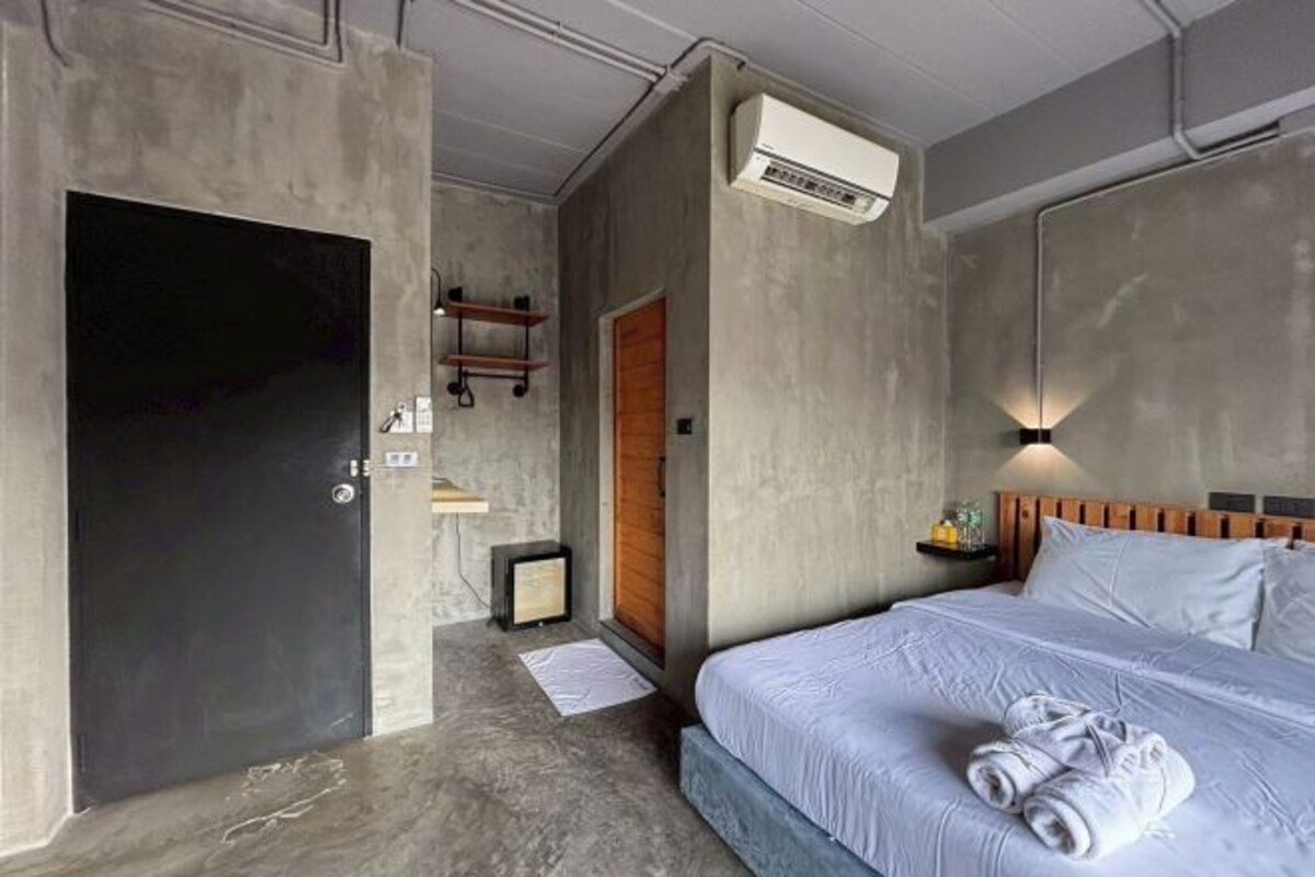 A bedroom with a bed, a wooden door, an air conditioning unit, a bedside table with a lamp, and a wall-mounted shelf.