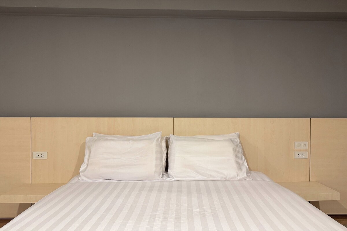 A bed with a striped bedspread and two pillows, a wooden headboard, and a nightstand with a power outlet.