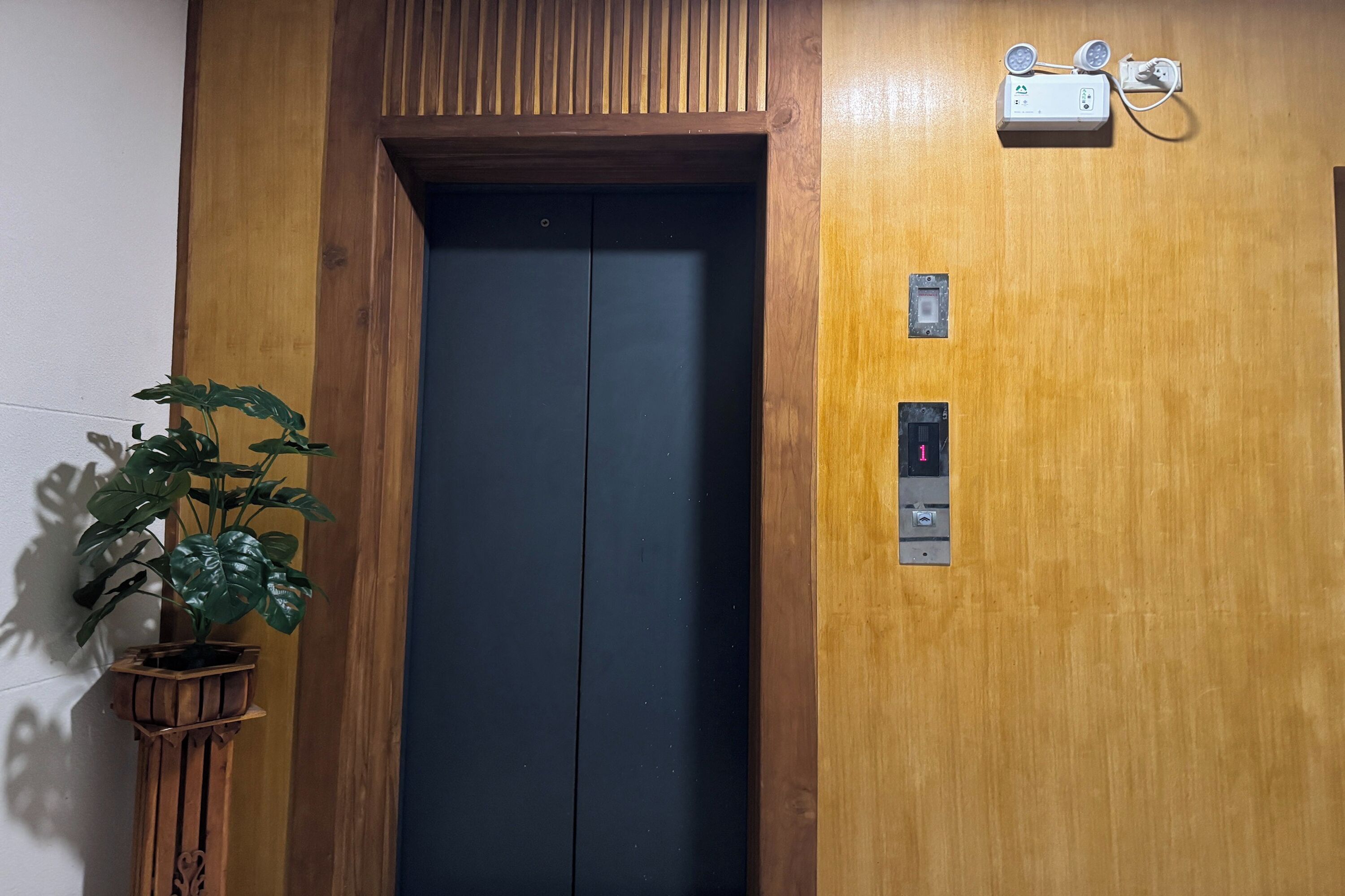 An elevator with a wooden finish, a potted plant beside it, and a wall-mounted intercom system.