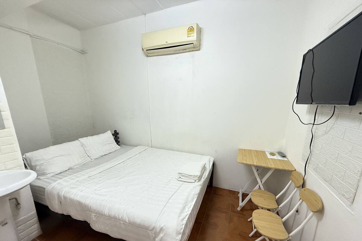 A small, clean room with a bed, a desk, a chair, an air conditioner, and a television.