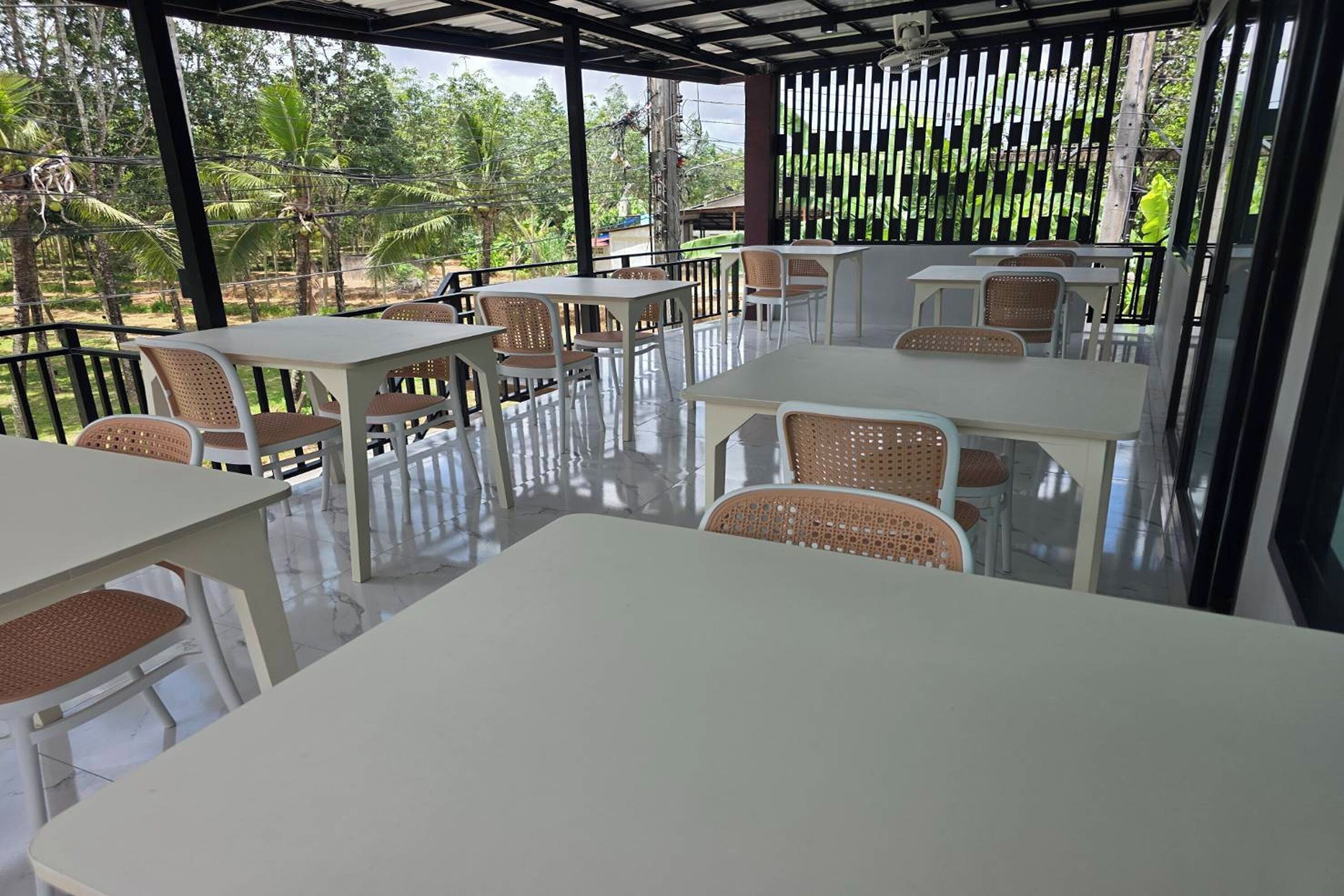 Un espacio de comedor al aire libre con mesas blancas y sillas de mimbre, rodeado por una cerca metálica y árboles.