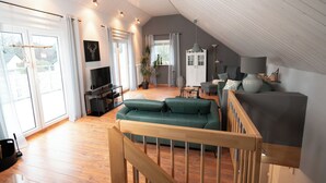 A spacious living room with a wooden staircase, a green sofa, a television, and a dining area with a table and chairs. - "Glücklich am Harz" dog-friendly vacation home with garden and fireplace (Goslar)