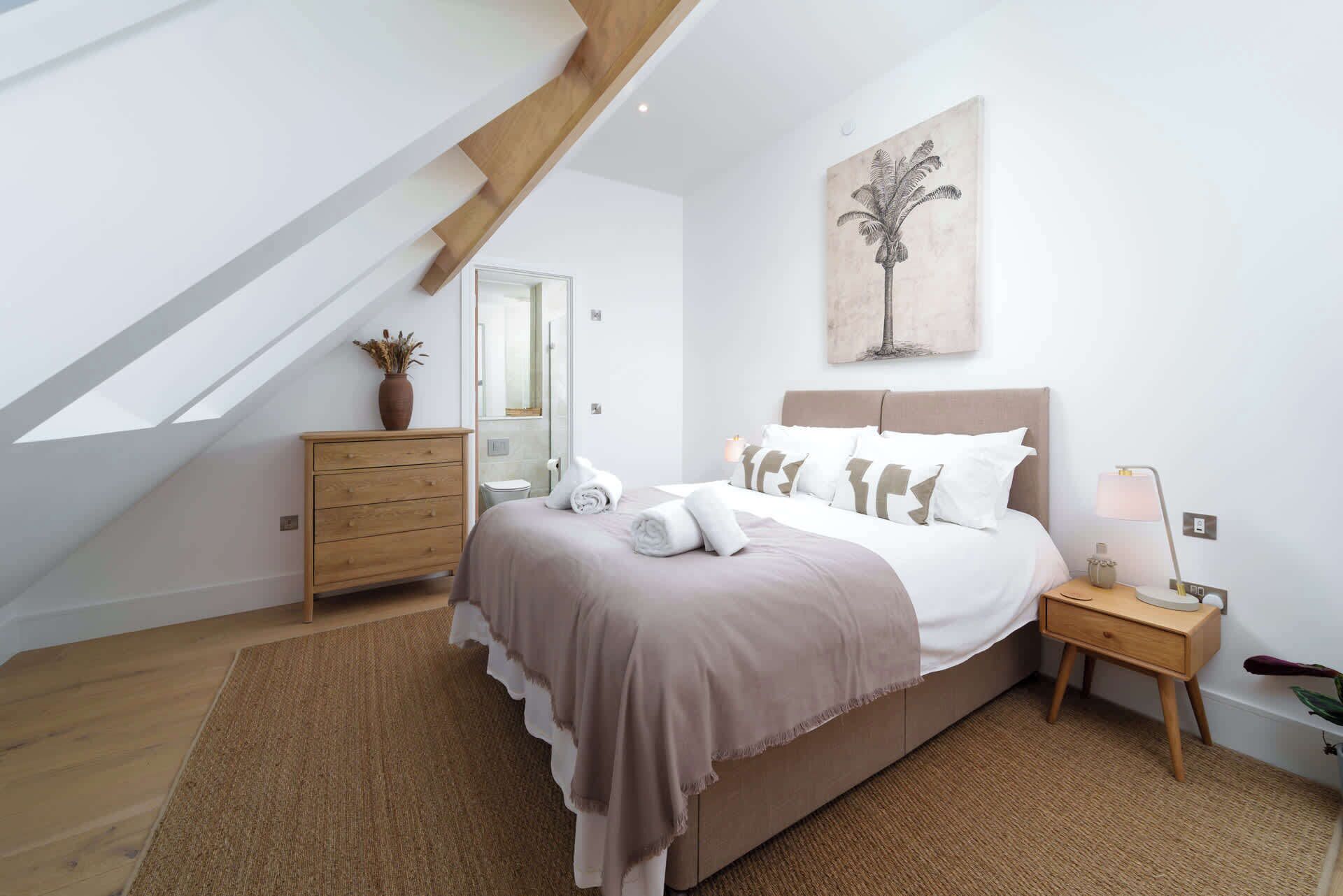 A bedroom with a sloped ceiling, a bed with a headboard, a nightstand, a lamp, a potted plant, and a painting on the wall.