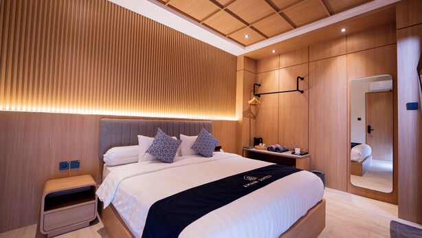 A modern hotel room with a large bed, a bedside table, a nightstand, a mirror, and a wooden panel wall. - Amani Suites Yogyakarta (Yogyakarta)