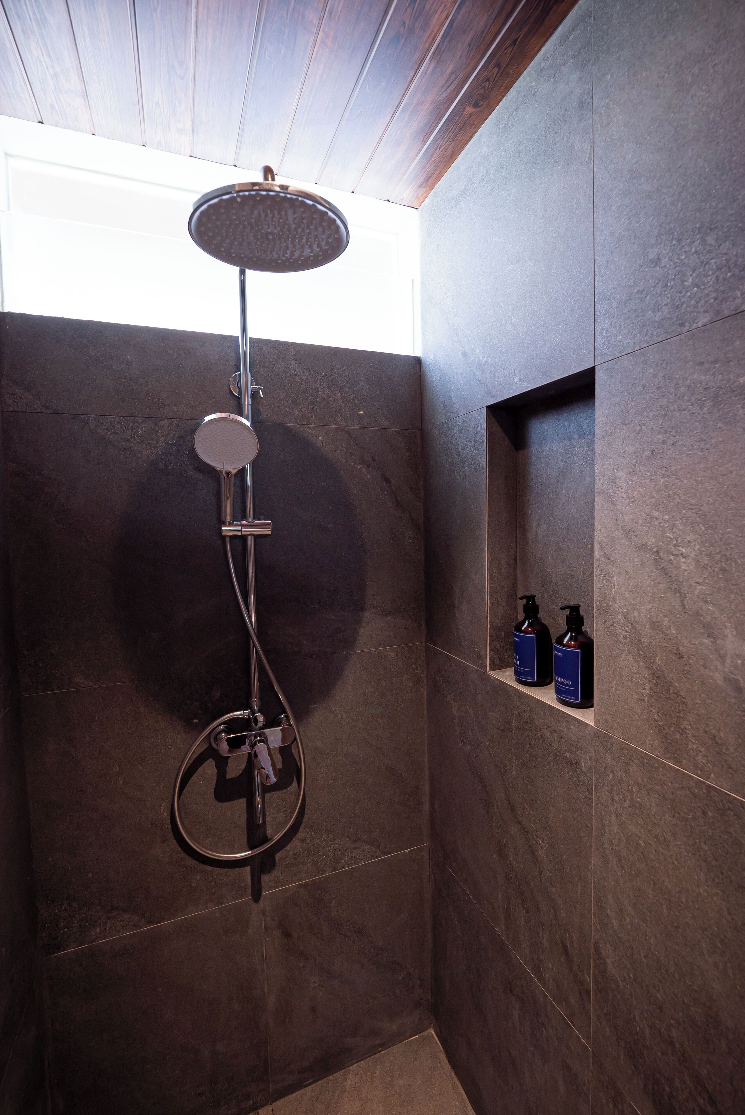 A modern shower with a rain showerhead, a handheld showerhead, and a handheld scrubber. There are two bottles of shampoo and conditioner on a shelf.