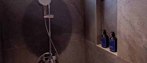 A modern shower with a rain showerhead, a handheld showerhead, and a handheld scrubber. There are two bottles of shampoo and conditioner on a shelf.