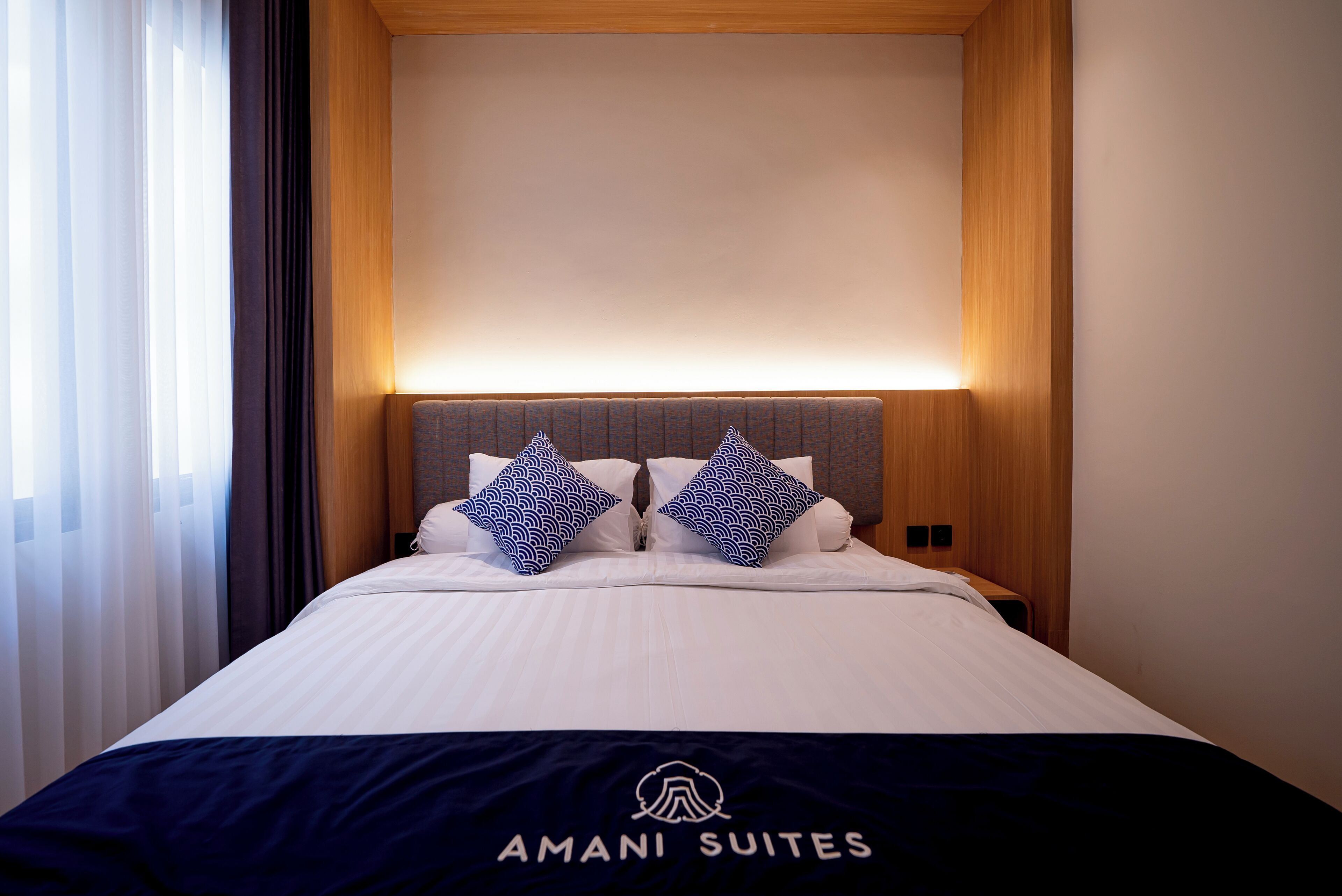 A hotel room with a bed, two patterned pillows, a dark blue bedspread, and a wooden headboard.