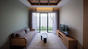 A modern living room with a sofa, a coffee table, a TV on a wooden stand, and a large window with curtains. - Amani Suites Yogyakarta (Yogyakarta)