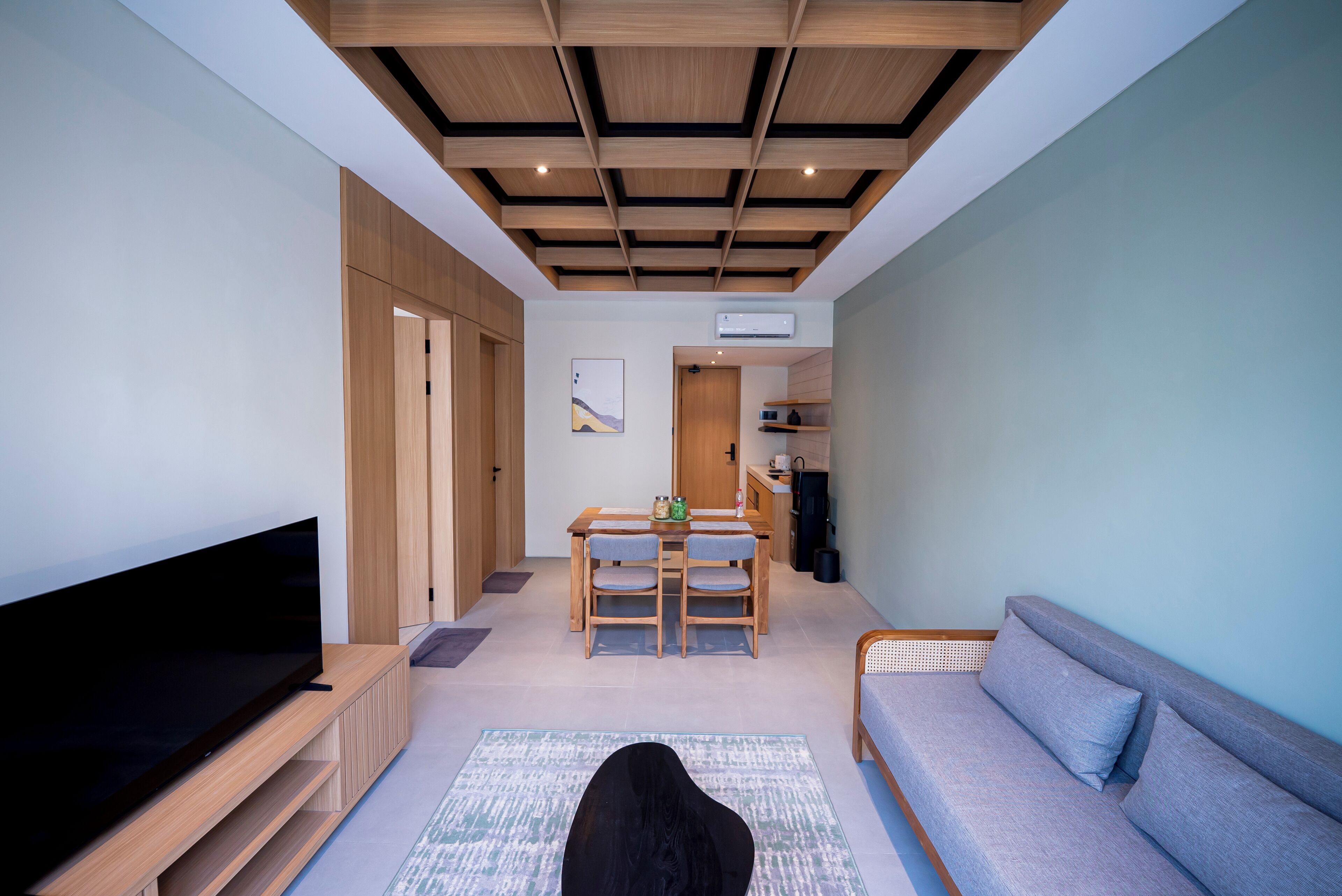 A modern living room with a flat-screen TV, a sofa, a dining area with a table and chairs, and a kitchen area visible in the background.