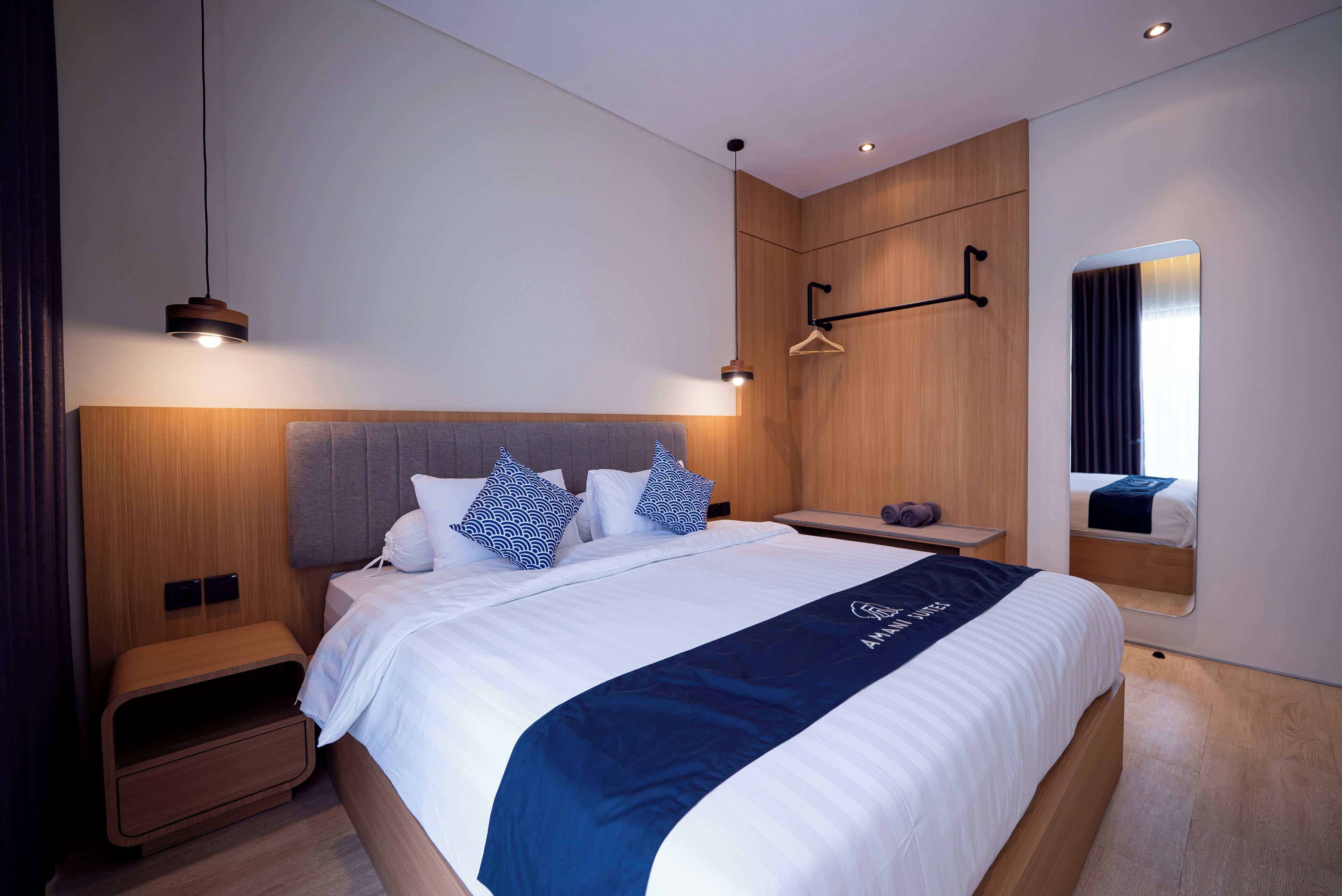 Una habitación de hotel moderna con una cama grande, cabecera de madera, mesitas de noche, un espejo y una lámpara colgante.