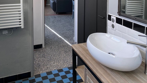A modern bathroom with a white sink, a mirror, and a towel rack. There is a checkered floor and a door leading to a living area with a couch and a plant. - Self-contained apartment in village house (Saint-André-de-Sangonis)