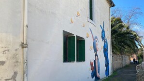 A narrow alley with a white building featuring a mural of a woman and a blue creature. - Self-contained apartment in village house (Saint-André-de-Sangonis)