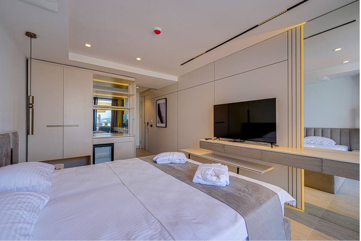 A modern hotel room with a large bed, a TV, a desk with a chair, and a minibar.
